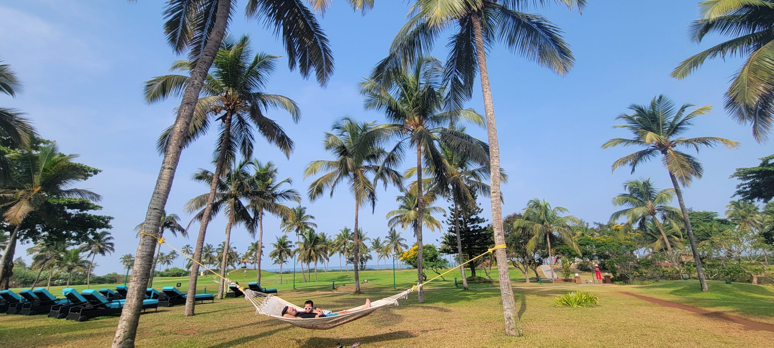 Caravela Beach Resort Goa family activities - Person in hammock amidst palm trees with lush greenery.