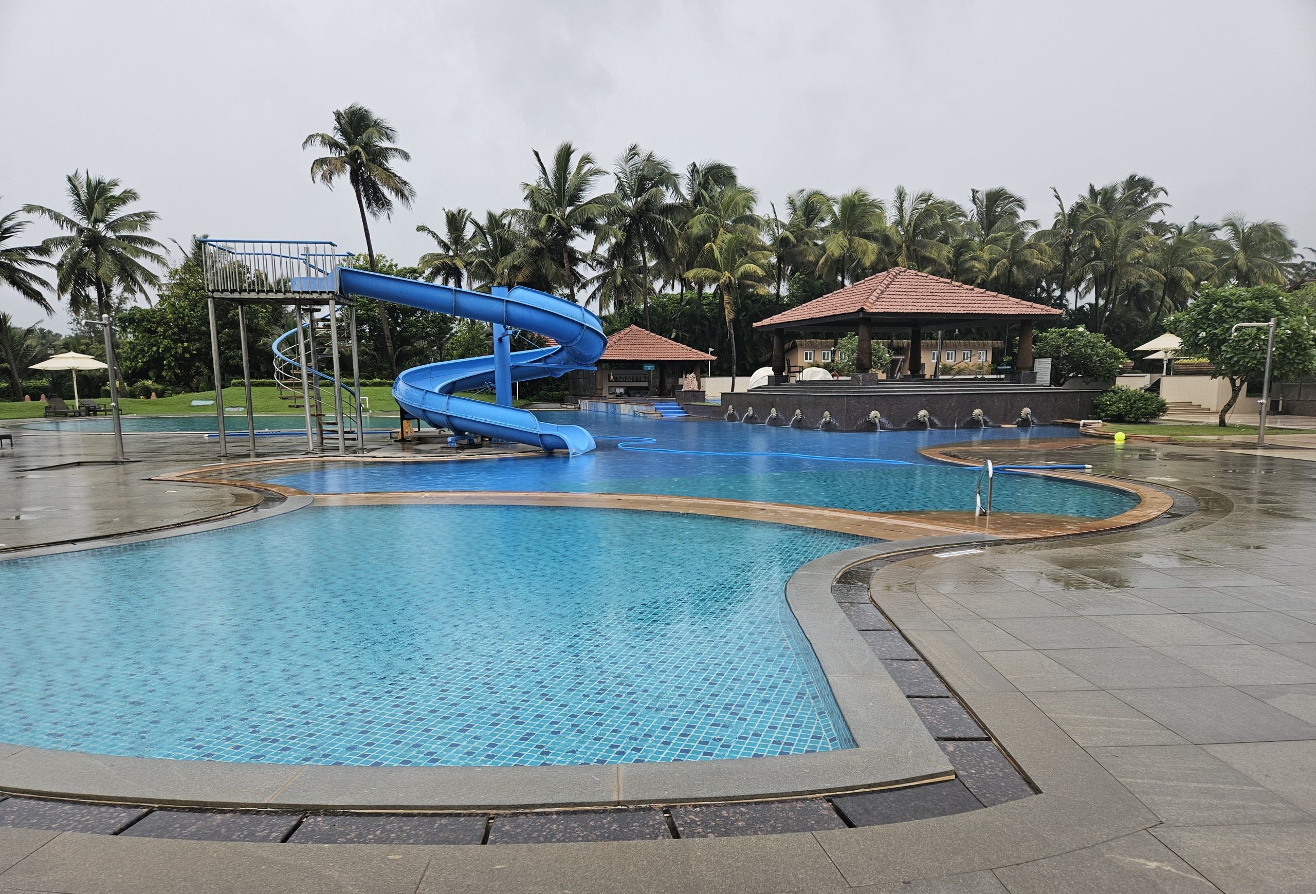 Kenilworth Resort & Spa Goa pool area - Large pool with blue water slide, surrounded by lush palm trees and greenery. Poolside bar with red-tiled roof in the background, capturing the resort’s serene and inviting environment.