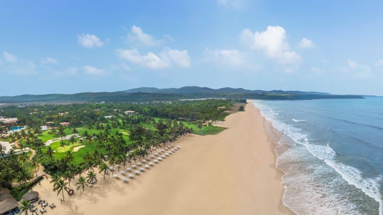 Panoramic view of St. Regis Goa Resort beach - Sandy shore with umbrellas and lounge chairs, lush greenery, and palm trees against a clear blue sky with hills in the background